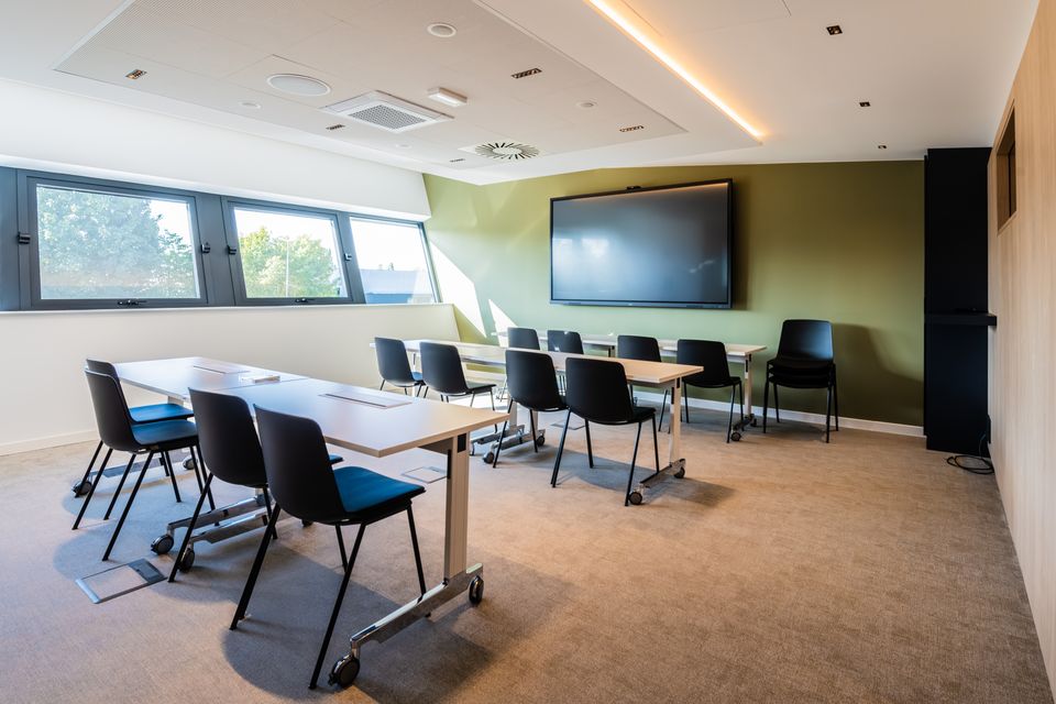 La salle de formation : tables et chaises face à un grand écran mural, éclairée par la lumière du jour.